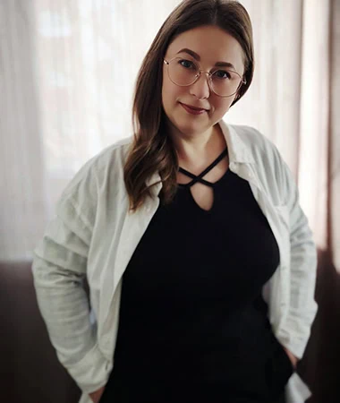 Portrait von Maryna Zakharova mit braunen Haaren, Brille und weißem Cardigan vor hellem Hintergrund, schwarzes Oberteil, direkter Blick in die Kamera